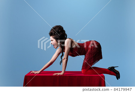 Portrait of young adorable woman in dark red, wine color evening dress posing on table with red cloth isolated on blue background. Portrait of young adorable woman in dark red, wine color evening dress posing on table with red cloth isolated on blue background. 84179947