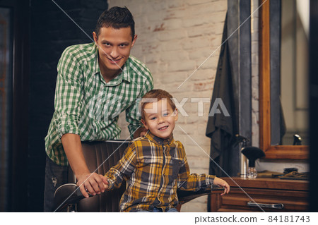 stylish little kid sitting on chair at barbershop with his young father on background stylish little kid sitting on chair at barbershop with his young father on background 84181743