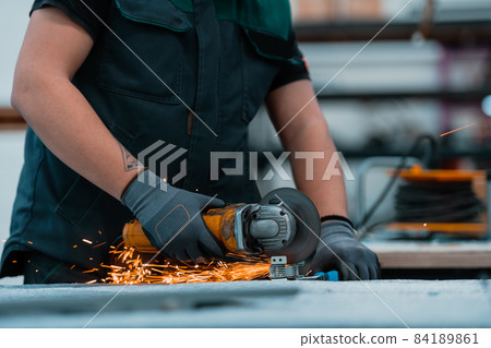 Heavy Industry Engineering Factory Interior with Industrial Worker Using Angle Grinder and Cutting a Metal Tube.He Wears a Mask on His Face Because of the Coronavirus Pandemic 84189861
