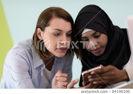 afro girl with a hijab and a European woman use a cell phone and laptop in their home office afro girl with a hijab and a European woman use a cell phone and laptop in their home office 84190100