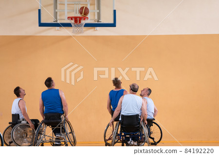 Disabled War veterans mixed race opposing basketball teams in wheelchairs photographed in action while playing an important match in a modern hall. Disabled War veterans mixed race opposing basketball teams in wheelchairs photographed in action while playing an important match in a modern hall. 84192220