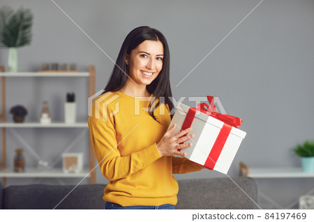 Happy girl with a gift box with a red ribbon sitting in the room. Surprise on Valentine's Day. 84197469