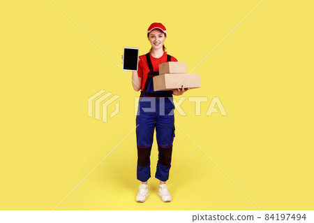 Full length portrait of courier woman holding parcels for delivery and showing tablet with blank screen for advertisement, wearing overalls and cap. Indoor studio shot isolated on yellow background. Full length portrait of courier woman holding parcels for delivery and showing tablet with blank screen for advertisement, wearing overalls and cap. Indoor studio shot isolated on yellow background. 84197494