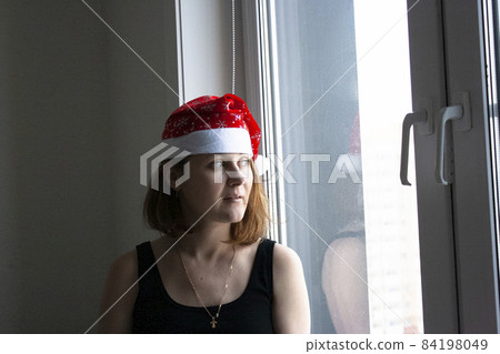 A young woman in a red Christmas cap in a white interior by the window. Christmas mood. A young woman in a red Christmas cap in a white interior by the window. Christmas mood. 84198049