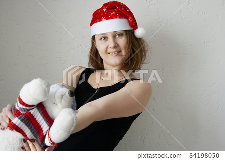 A young woman in a red Christmas cap with a stuffed husky dog toy in her hands in a white interior. 84198050