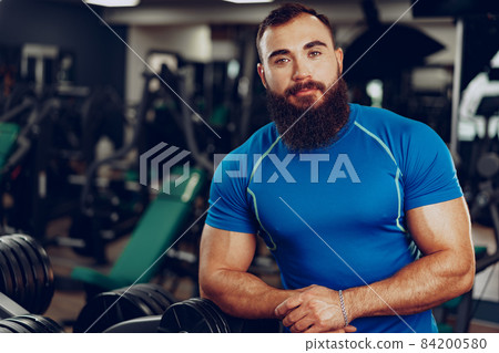Bearded young man bodybuilder in blue t-shirt standing in gym Bearded young man bodybuilder in blue t-shirt standing in gym 84200580