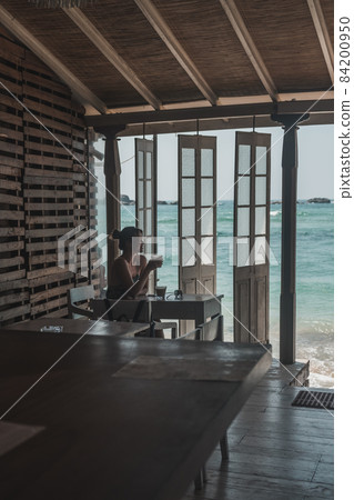 Girl sitting and drinking coffee in a wooden cafe overlooking the ocean. Blue crystal clear water in the ocean. Lovely view from the coffee shop 84200950