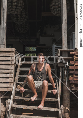 Young handsome caucasian man with a sleeve tattoo sits on the wooden stairs of a cozy era friendly cafe in Asian. Overlooks the ocean. 84200983