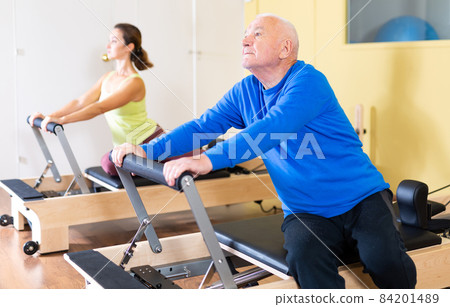 Mature man practicing Pilates system on reformer supervised in sport gym Mature man practicing Pilates system on reformer supervised in sport gym 84201489
