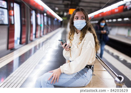 Girl in a protective mask sitting on a bench in the subway with a mobile phone waiting for a train 84201629