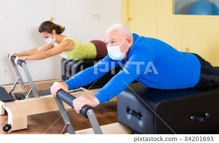 Mature man in protective mask practicing Pilates system on reformer supervised in sport gym 84201693