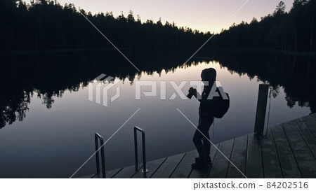 Silhouette traveler photographing scenic view in forest, river. Wood pier. One woman shooting nice dark magic night look. Girl take photo on camera. Silhouette traveler photographing scenic view in forest, river. Wood pier. One woman shooting nice dark magic night look. Girl take photo on camera. 84202516