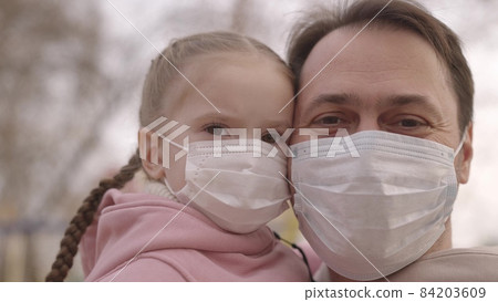 father with little child girl in medical mask smiles, dad walks with the kid on the playground, hugs his beloved baby holding in his arms, happy family, father's day, man with daughter close-up face 84203609