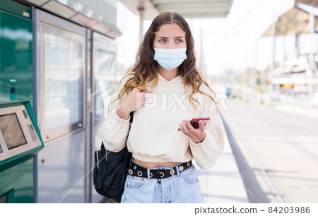 Woman in mask holding smartphone while waiting for tram Woman in mask holding smartphone while waiting for tram 84203986