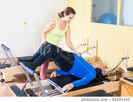 Woman pilates instructor helping aged man doing exercises on reformer Woman pilates instructor helping aged man doing exercises on reformer 84205143