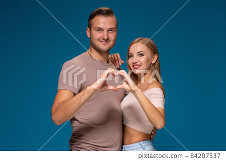 Happy couple dressed casual clothes, making heart shape from fingers, studio portrait on blue background 84207537