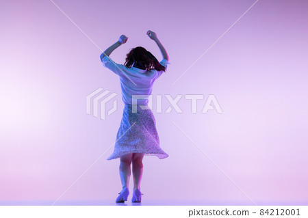 Back view of young excited woman in vintage retro style clothes dancing isolated on lilac color background in neon light 84212001