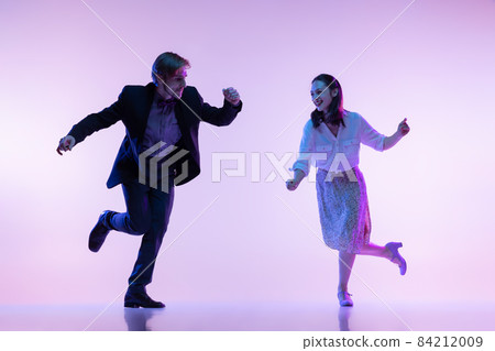 Dynamic portrait of young dancers, man and woman in vintage style outfits dancing swing isolated on lilac color background in neon light 84212009