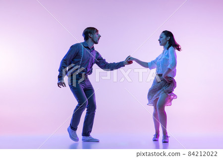 Studio shot of young man and woman in vintage retro style outfits dancing lindy hop isolated on lilac color background in neon light Studio shot of young man and woman in vintage retro style outfits dancing lindy hop isolated on lilac color background in neon light 84212022