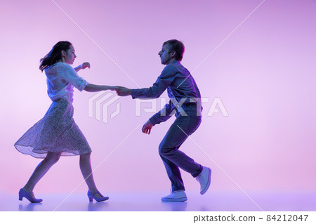 Two young graceful dancers, man and woman in vintage style outfits dancing rock-and-roll isolated on lilac color background in neon light Two young graceful dancers, man and woman in vintage style outfits dancing rock-and-roll isolated on lilac color background in neon light 84212047