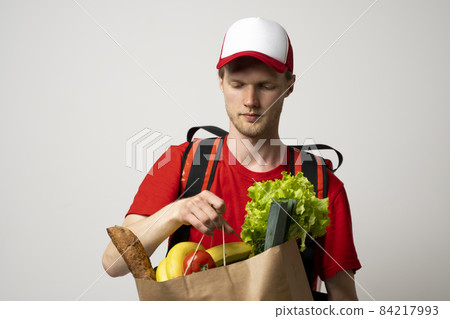Grocery delivery courier man in a red cap and t-shirt with a thermal bag on a shoulders holds paper bag with food on white studio background. Food delivery service, online shopping. 84217993