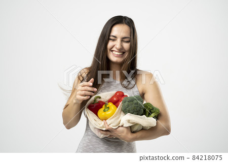 Smiling brunette girl is holding mesh shopping bag with vegetables, greens without plastic bags. Zero waste, plastic free Eco friendly concept. Sustainable lifestyle. 84218075