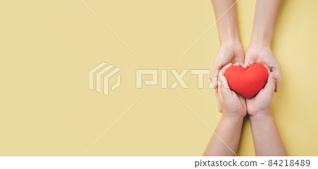 Mother and son hand holding red heart on yellow background, heart health insurance, charity volunteer donation, CSR responsibility, world heart day, world health day, family day, saving lives. Mother and son hand holding red heart on yellow background, heart health insurance, charity volunteer donation, CSR responsibility, world heart day, world health day, family day, saving lives. 84218489
