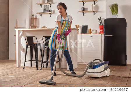 Young woman using vacuum cleaner in home kitchen floor, doing cleaning duties and chores, meticulous interior. Young woman using vacuum cleaner in home kitchen floor, doing cleaning duties and chores, meticulous interior. 84219657