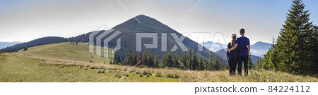 Back view of couple athletic boy and blond girl standing together embraced enjoying breathtaking view of magnificent Carpathian mountains, covered with ever-green forest on spring morning. Back view of couple athletic boy and blond girl standing together embraced enjoying breathtaking view of magnificent Carpathian mountains, covered with ever-green forest on spring morning. 84224112