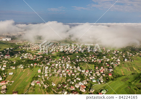Aerial view of white clouds above a town or village with rows of buildings and curvy streets between green fields in summer. Countryside landscape from above. Aerial view of white clouds above a town or village with rows of buildings and curvy streets between green fields in summer. Countryside landscape from above. 84224165