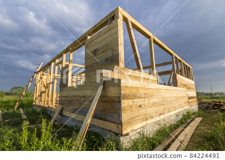 Unfinished ecological house under construction in green field on blue sky background. Trenches filled with cement and wooden walls with window openings of future comfortable cottage in quiet area. 84224491