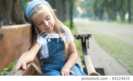 Small tired child girl sitting on a bench with closed eyes resting in summer park. 84224635