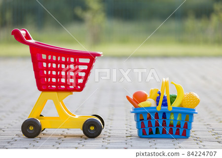 Shopping pushcart and a basket with toy fruits and vegetables. Bright plastic colorful toys for children outdoors on sunny summer day. Shopping pushcart and a basket with toy fruits and vegetables. Bright plastic colorful toys for children outdoors on sunny summer day. 84224707