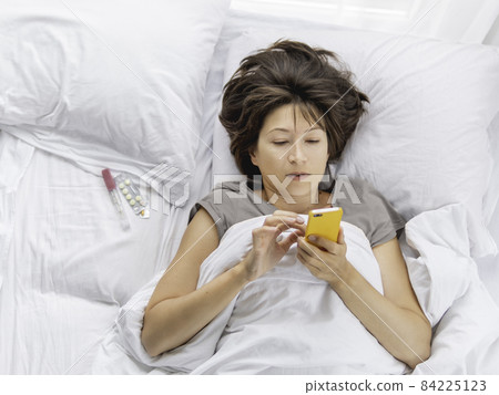 Top view on sick woman lying in bed with smartphone. Thermometer and pills on white bed linen. Internet surfing. 84225123