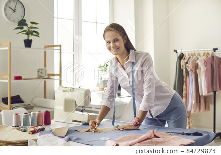 Dressmaker, seamstress or tailor working in modern atelier. Happy woman with pencil and cutting pattern marking fabric standing at table with measuring tape, thread, needles, pins and sewing machine 84229873