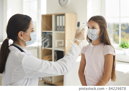 Nurse or doctor using modern digital infrared thermometer and pointing it at kid's forehead. Child who's wearing face mask gets body temperature checked at hospital. Covid 19, health, safety concepts 84229878