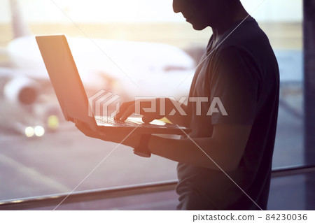 A man is working on a laptop at the airport while waiting to board the plane A man is working on a laptop at the airport while waiting to board the plane 84230036