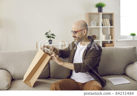 Satisfied adult male customer who received package from online store unpacks his order. Man sitting on sofa near consignment note and pulls out cardboard box delivered to him by delivery service. Satisfied adult male customer who received package from online store unpacks his order. Man sitting on sofa near consignment note and pulls out cardboard box delivered to him by delivery service. 84231484