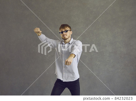 Studio portrait of happy redhead young man in thug life glasses dancing gangnam style. Cheerful guy with red hair wearing funny sunglasses having fun and dancing to music isolated on grey background 84232023