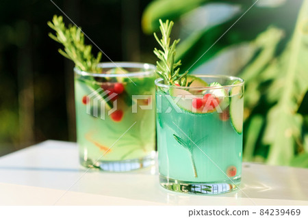 Two glasses with green lemonade made from lemon rosemary and mint in glasses on table at the terrace. Cold refreshing beverage for hot summer day. Green monstera leaves on background. 84239469