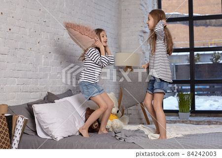 The image of two little sisters sitting on the bed in the room. They using pillow to fight each other The image of two little sisters sitting on the bed in the room. They using pillow to fight each other 84242003