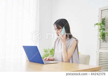 A female living room who looks at a computer and makes a phone call A female living room who looks at a computer and makes a phone call 84242376