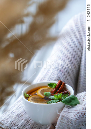 Female hands on sweater holding tea with lemon, cinnamon sticks and mint leaves. Hot drink. Healthy melissa tea natural organic aromatic drink in cup. Autumn winter warming drink. Cozy home 84242536