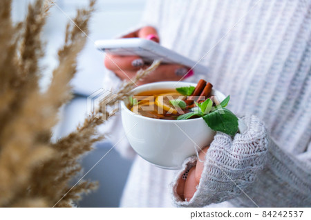 Female hands on sweater using phone and drinking tea with lemon, cinnamon sticks and mint leaves. Hot drink. Healthy melissa tea natural organic aromatic drink in cup. Autumn winter warming drink. Female hands on sweater using phone and drinking tea with lemon, cinnamon sticks and mint leaves. Hot drink. Healthy melissa tea natural organic aromatic drink in cup. Autumn winter warming drink. 84242537