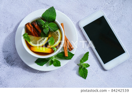 Black and green tea with lemon, cinnamon sticks and mint leaves on table with mobile phone. Social media. Healthy melissa tea natural organic aromatic drink in cup. Summer, Autumn winter warming Black and green tea with lemon, cinnamon sticks and mint leaves on table with mobile phone. Social media. Healthy melissa tea natural organic aromatic drink in cup. Summer, Autumn winter warming 84242539