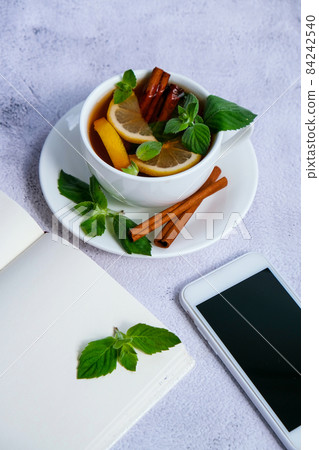 Black and green tea with lemon, cinnamon sticks and mint leaves on table with mobile phone. Social media. Healthy melissa tea natural organic aromatic drink in cup. Summer, Autumn winter warming Black and green tea with lemon, cinnamon sticks and mint leaves on table with mobile phone. Social media. Healthy melissa tea natural organic aromatic drink in cup. Summer, Autumn winter warming 84242540