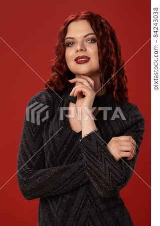 Beautiful sexy woman is posing in a knitted dress on a red background. Evening makeup. Long, healthy, red curly hair. Beautiful sexy woman is posing in a knitted dress on a red background. Evening makeup. Long, healthy, red curly hair. 84243038