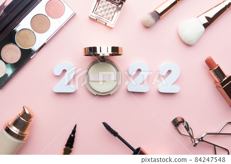 2022 Beauty cosmetic makeup products trends concept. Top view of 2022 white number with powder, lipstick, foundation, eyeshadow, brush, eyeliner and mascara on pink background. 2022 Beauty cosmetic makeup products trends concept. Top view of 2022 white number with powder, lipstick, foundation, eyeshadow, brush, eyeliner and mascara on pink background. 84247548