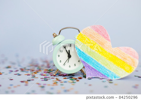Alarm clock showing five minutes to twelve with milticolored sequins and lgtb flag in the form of a heart 84250296