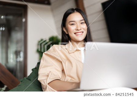 Smiling asian woman using laptop computer at home and looking happy at camera 84251545